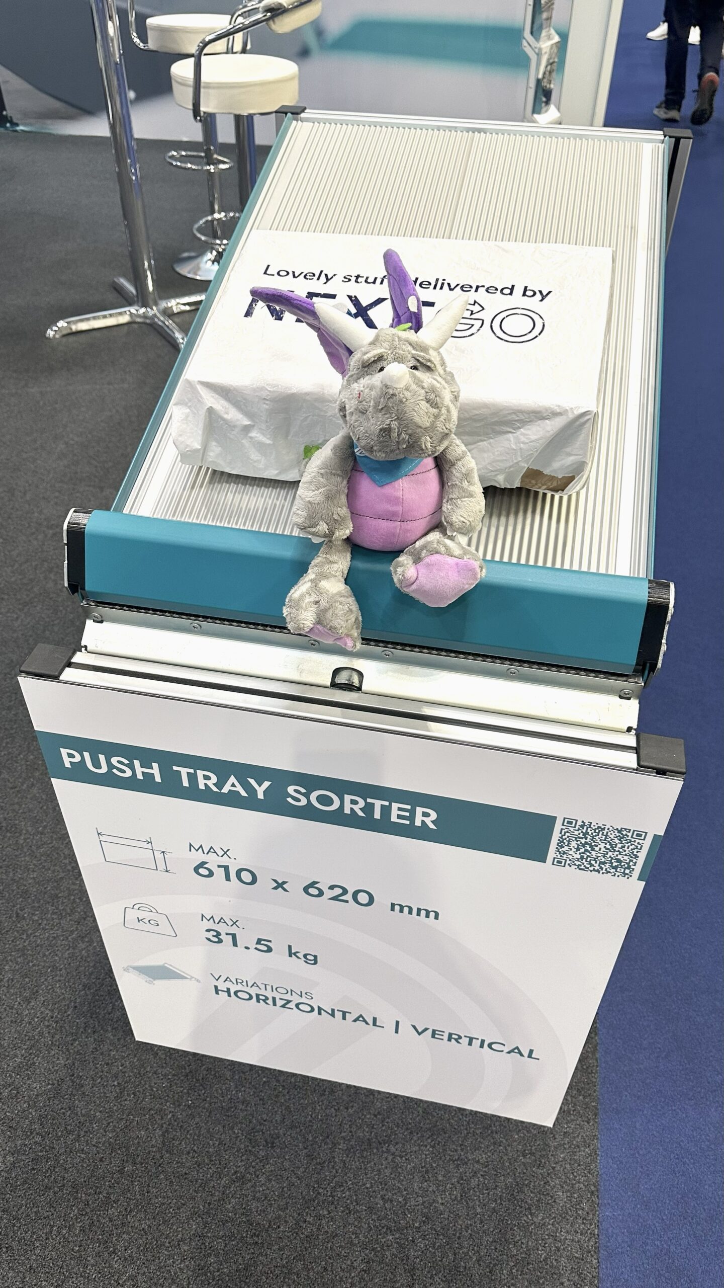 A gray and purple plush dragon sits on a conveyor belt in front of a sign labeled Push Tray Sorter, with a parcel behind it. The setting appears to be an exhibition or trade show.