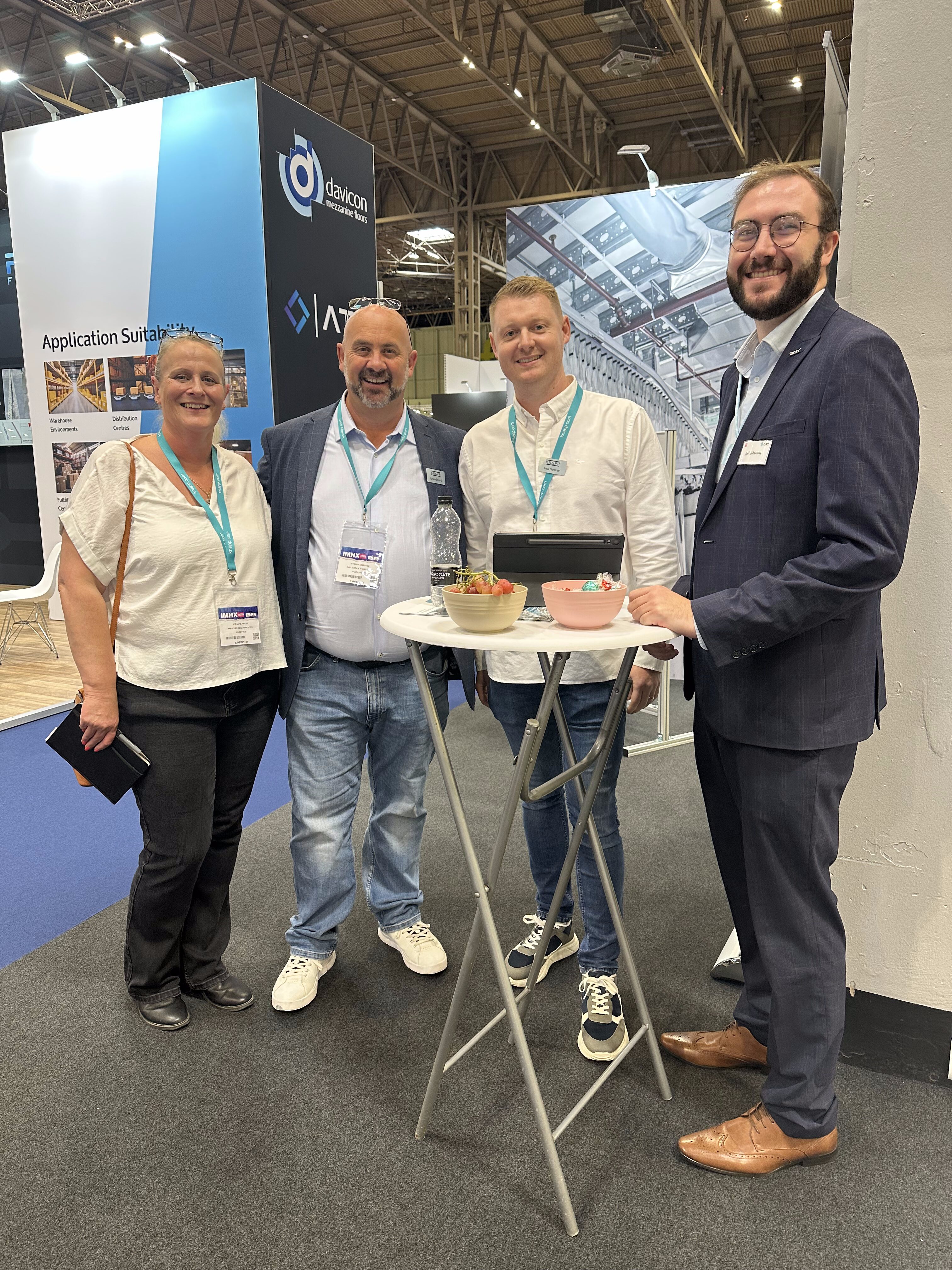 Four people are standing and smiling around a tall round table at a trade show or conference, with badges and lanyards visible. The table holds snacks, drinks, and a tablet. Exhibition booths are in the background.
