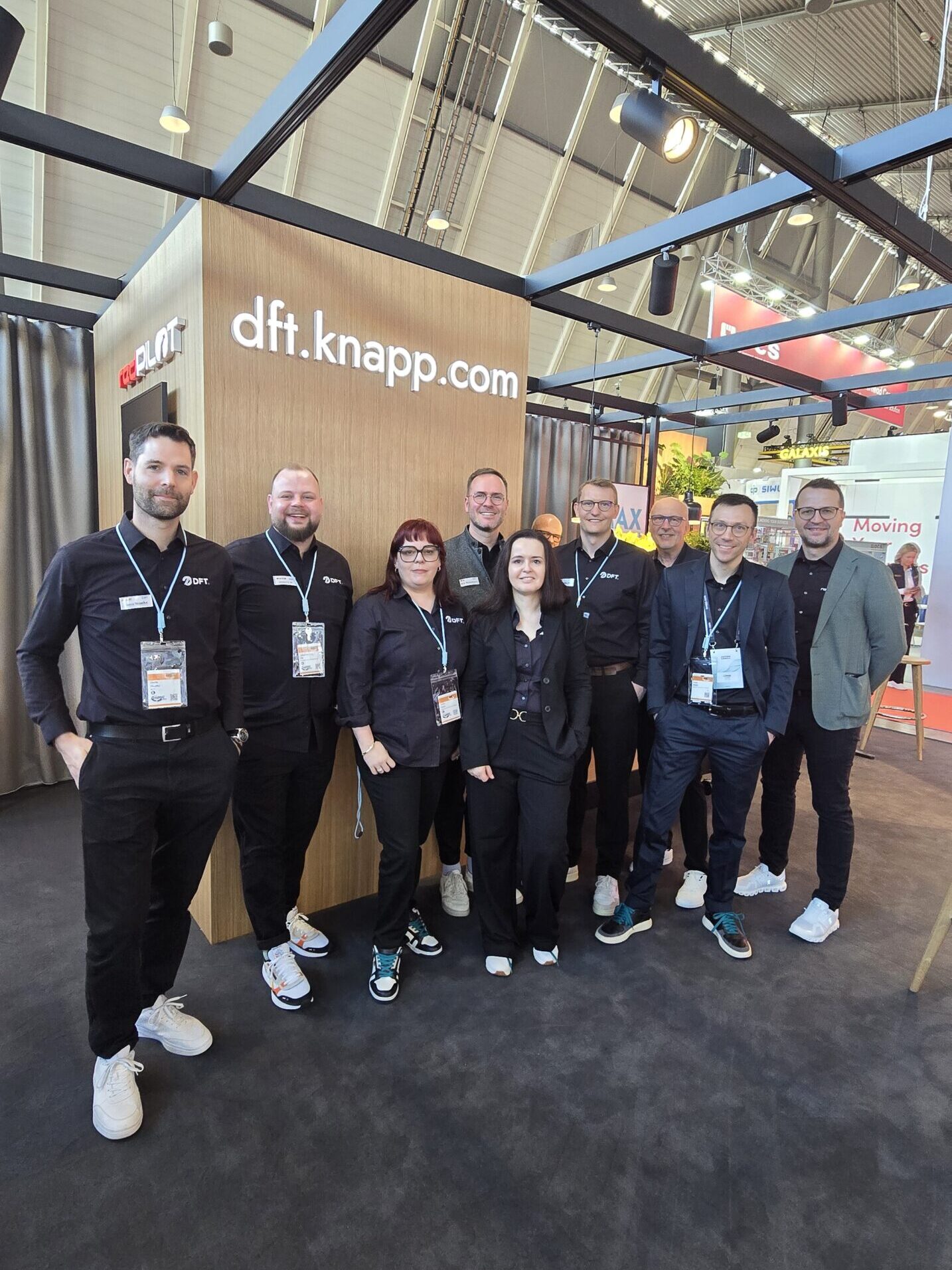 Eine Gruppe von zehn Personen in schwarzer Gesch&auml;ftskleidung steht l&auml;chelnd vor einem h&ouml;lzernen Stand, der die Website dft.knapp.com auf einer Messe oder Ausstellung zeigt.
