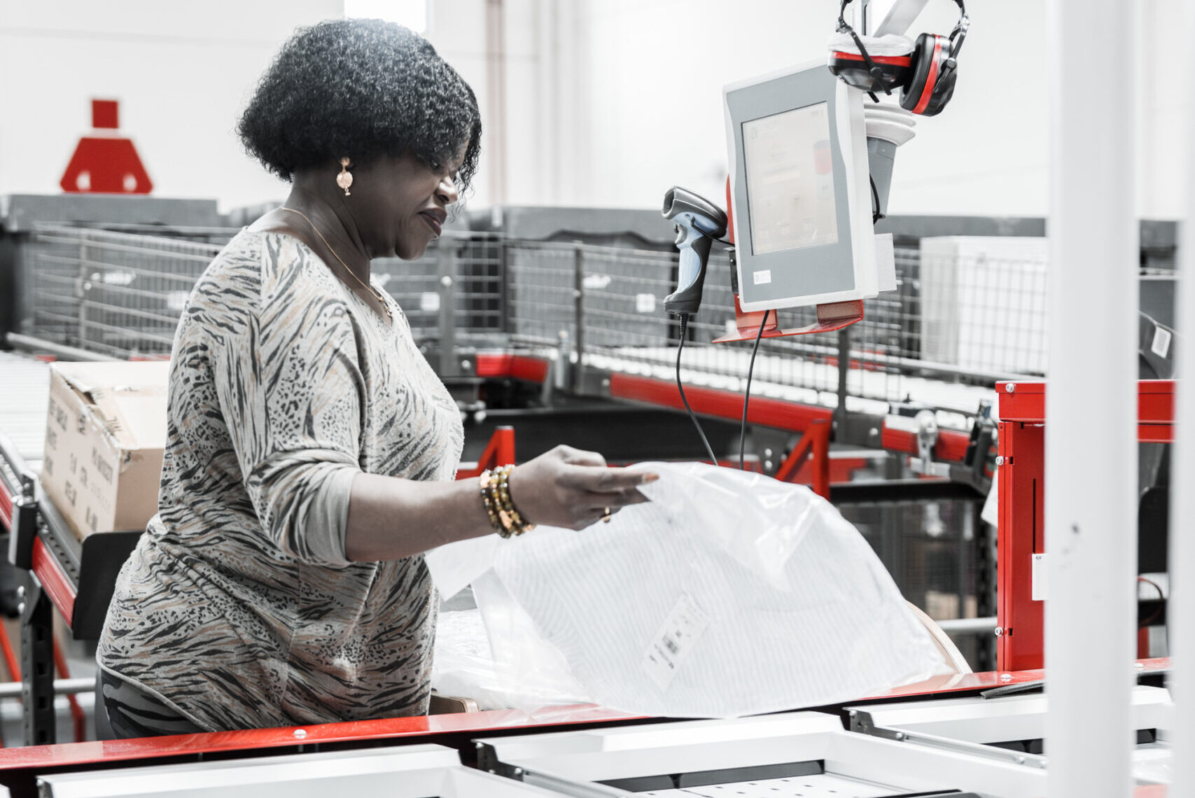 A woman works at a packaging station in a warehouse for the apparel industry, handling products and using a touchscreen monitor, with red machinery and shelves in the background.