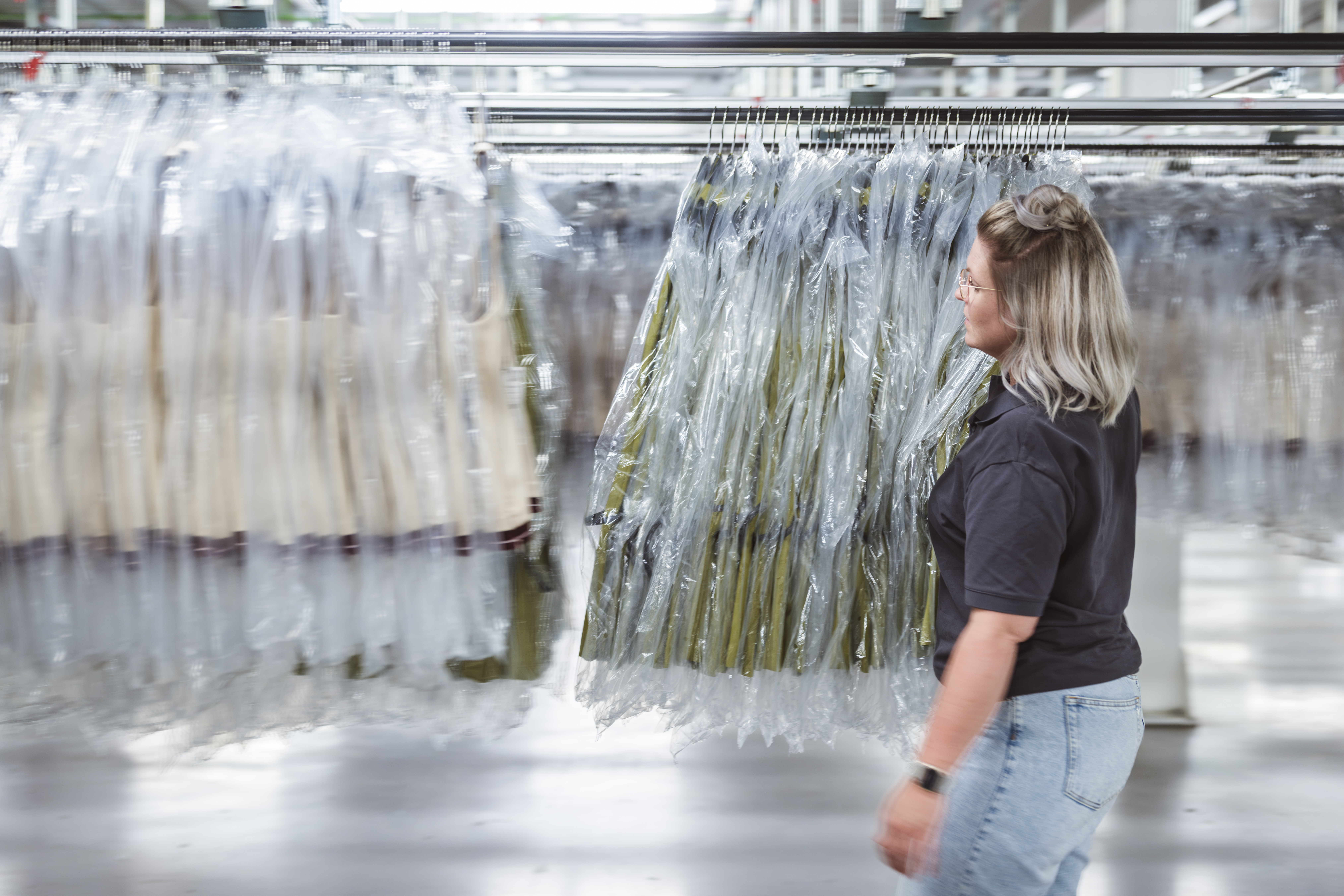 A woman in a black shirt and jeans examines rows of clothing covered in plastic hanging on racks in a large, brightly lit apparel industry facility.