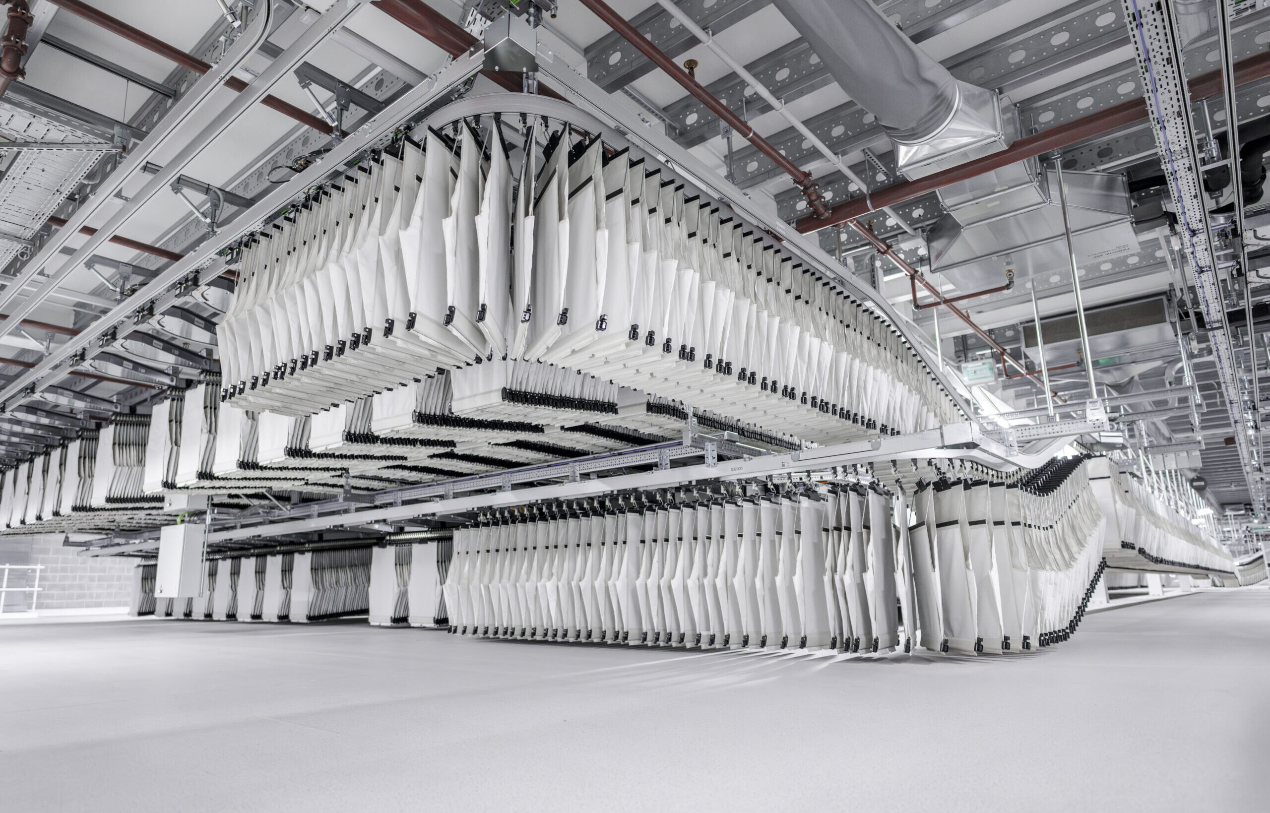 Ein großes Textilfördersystem mit zahlreichen weißen Stoffstücken, die an Schienen in einer geräumigen Industrieumgebung aufgehängt sind. Das System ist in Reihen angeordnet und bildet vor dem Hintergrund einer metallischen Decke ein komplexes, geordnetes Muster.