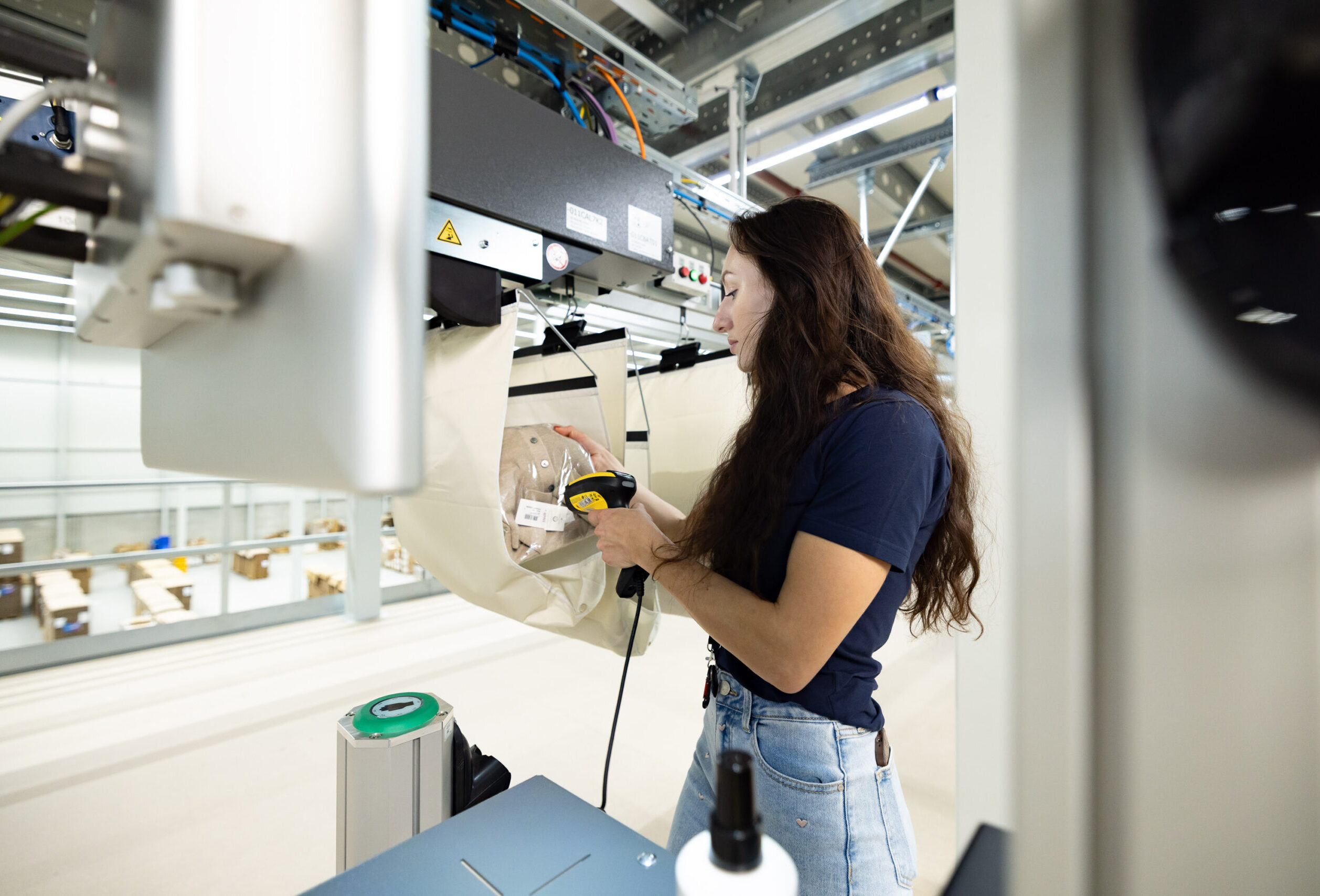 A woman with long brown hair uses a handheld scanner to scan a package at a modern logistics facility, surrounded by machinery, Pocket Sorter systems, and shelves.
