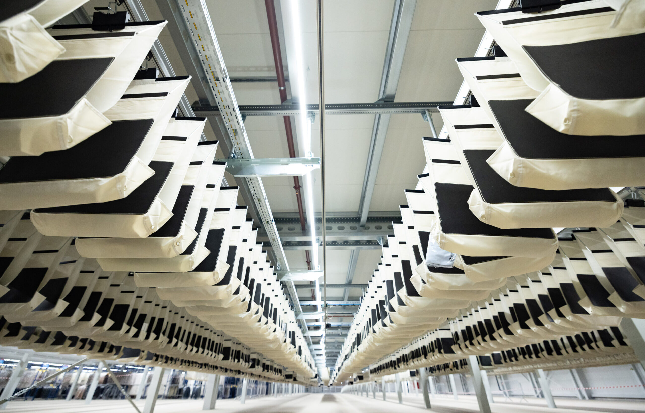 Blick aus geringer Höhe auf Reihen großer weißer und schwarzer EcoPocket-Stoffbeutel, die an Metallschienen in einer modernen Industrieanlage aufgehängt sind und einen symmetrischen, tunnelartigen Effekt erzeugen.