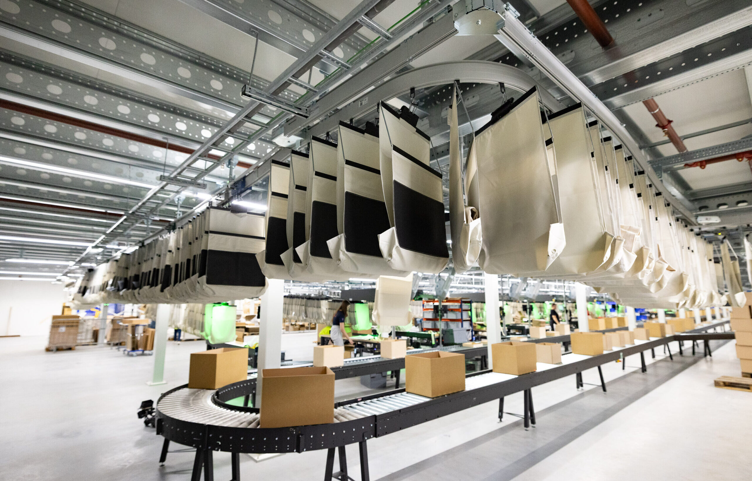 Ein Fördersystem in einem Lager, an dem leere Stoffsäcke an Haken von der Decke hängen und Kartons für die Ersatzteillogistik auf dem Förderband transportiert werden. Die Umgebung ist hell beleuchtet und übersichtlich gestaltet.