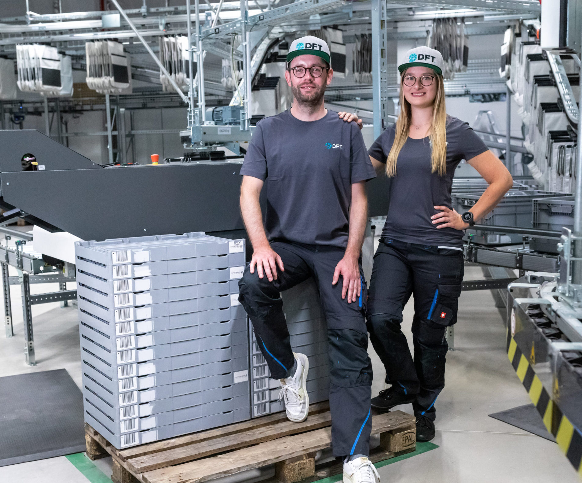 Zwei Personen in passender Arbeitskleidung und Helmen stehen in einer Industrieanlage. Einer sitzt auf einem Stapel Plastikkisten auf einer Palette, während der andere steht. Beide lächeln. Im Hintergrund sind Förderbänder und Maschinen zu sehen.