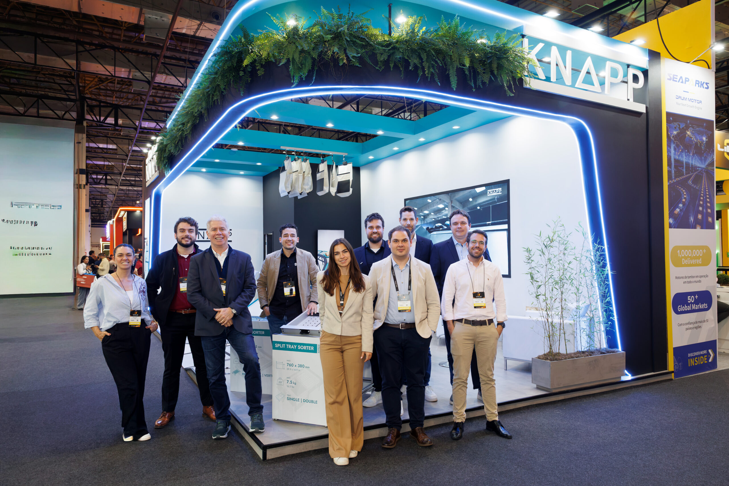 Eine Gruppe von zehn Personen steht l&auml;chelnd vor einem eleganten Messestand mit der Aufschrift "KNAPP", umgeben von Pflanzen und moderner Beleuchtung.