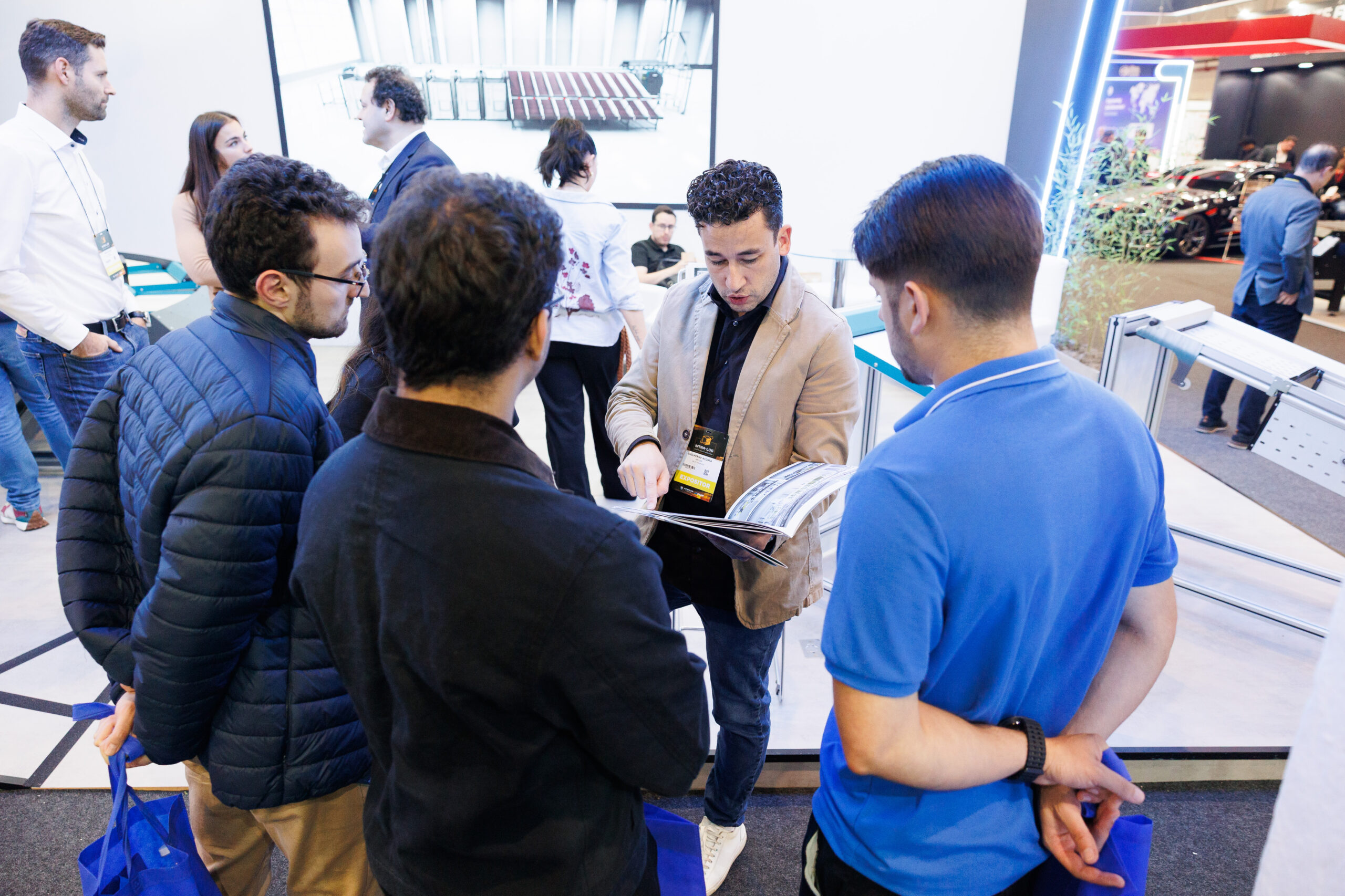 Eine Gruppe von Personen steht an einem Stand, w&auml;hrend ein Mann in einem beigen Blazer Informationen in einer Brosch&uuml;re zeigt und erkl&auml;rt.