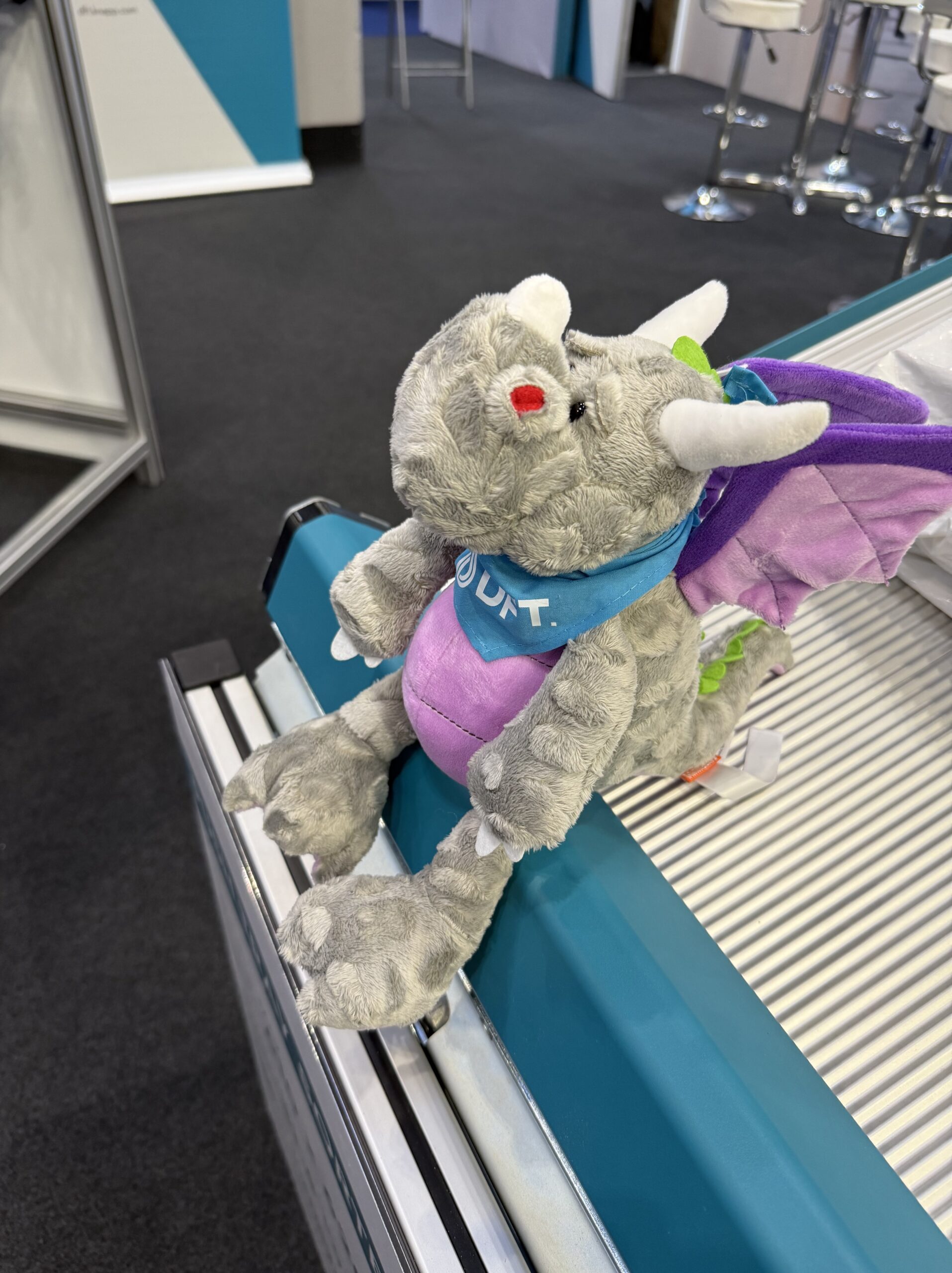 A small gray plush dragon with purple wings and a blue UKT bandana sits on the edge of a metallic, ridged surface in an indoor setting with dark carpeting.