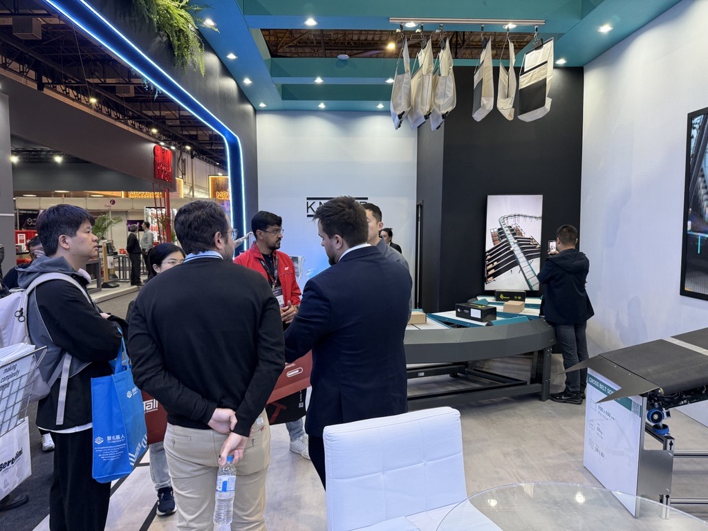 Eine Gruppe von Fachleuten diskutiert an einem Messestand, w&auml;hrend im Hintergrund ein Monitor und ein F&ouml;rderbandsystem zu sehen sind.