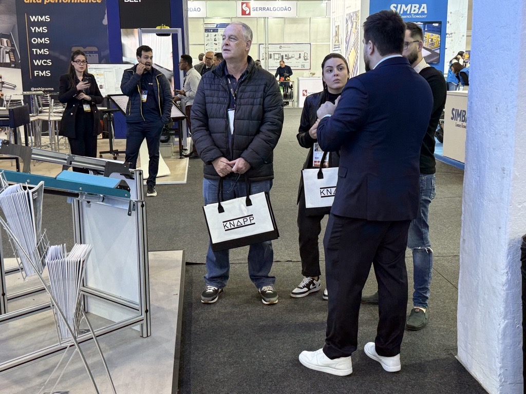Eine Gruppe von Fachleuten steht an einem Messestand und diskutiert, w&auml;hrend einige von ihnen Tragetaschen der Marke KNAPP halten.