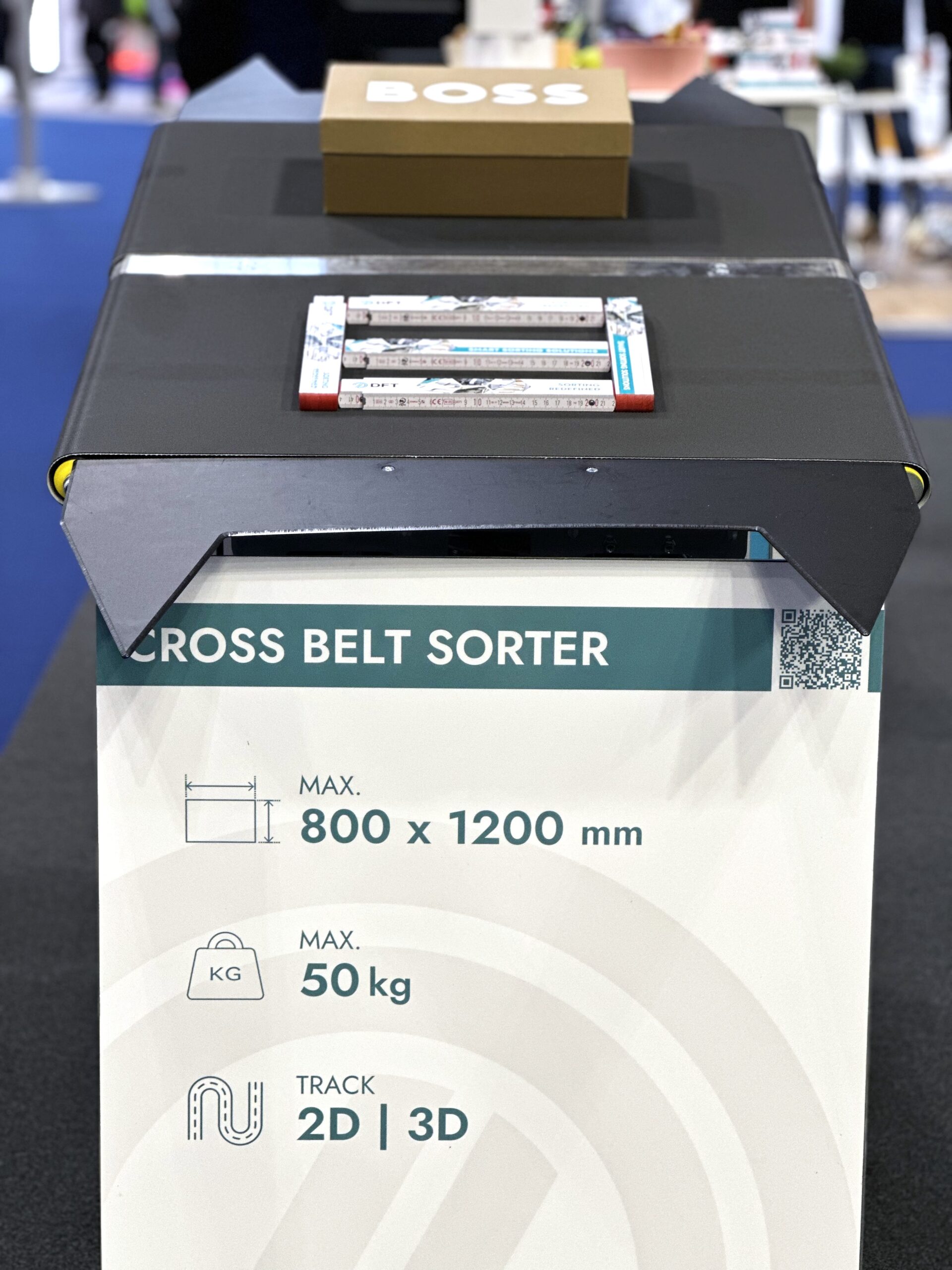 A cross belt sorter display shows a package and books on a conveyor. The sign lists specifications: max size 800 x 1200 mm, max weight 50 kg, and 2D/3D track options. The background is a blurred indoor setting.