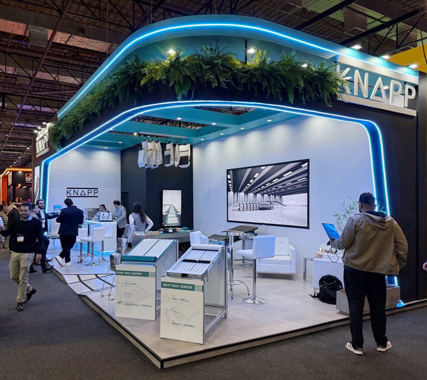 Ein moderner Messestand der Firma KNAPP mit ansprechender Beleuchtung, M&ouml;beln und ausgestellten Produkten, w&auml;hrend Besucher den Stand erkunden und interagieren.