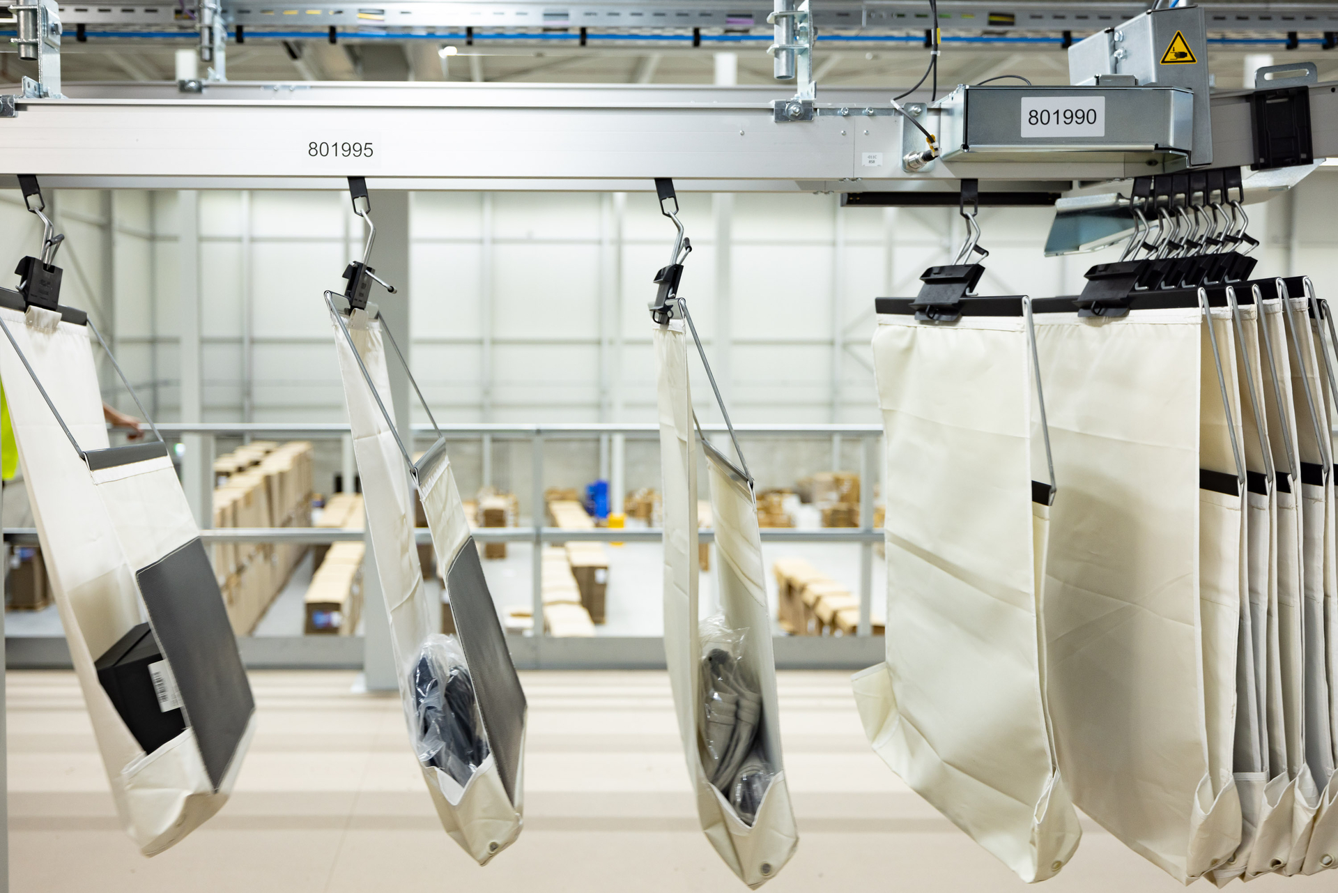 Large white bags with black trim hang on a conveyor system in a warehouse, each holding packaged items. Shelves and boxes are visible in the out-of-focus background.
