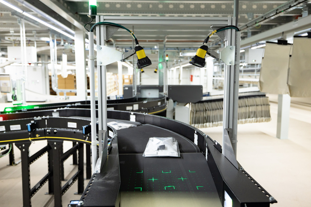 Automatisiertes Fördersystem in einem Lager, in dem Pakete unter Scannern und Kameras hindurchlaufen, von grünem Licht beleuchtet und von Industrieregalen und Geräten umgeben.