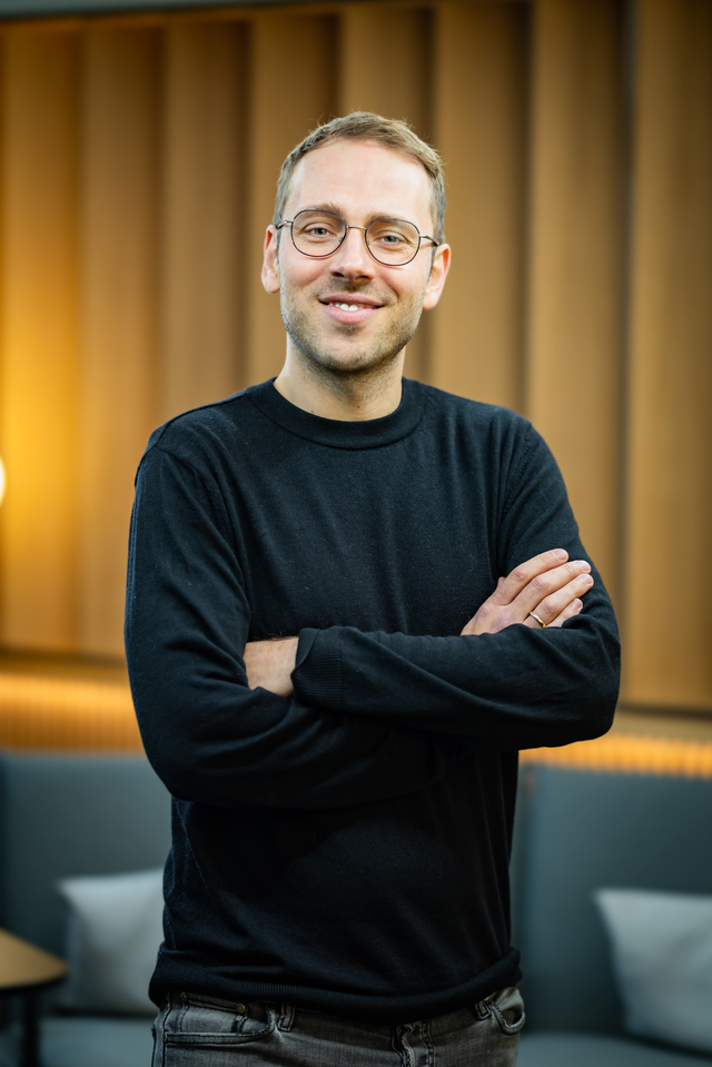 Portraitfoto Pascal Rehm: Ein Mann mit Brille steht l&auml;chelnd mit verschr&auml;nkten Armen vor einer modernen, hellen Hintergrundkulisse.