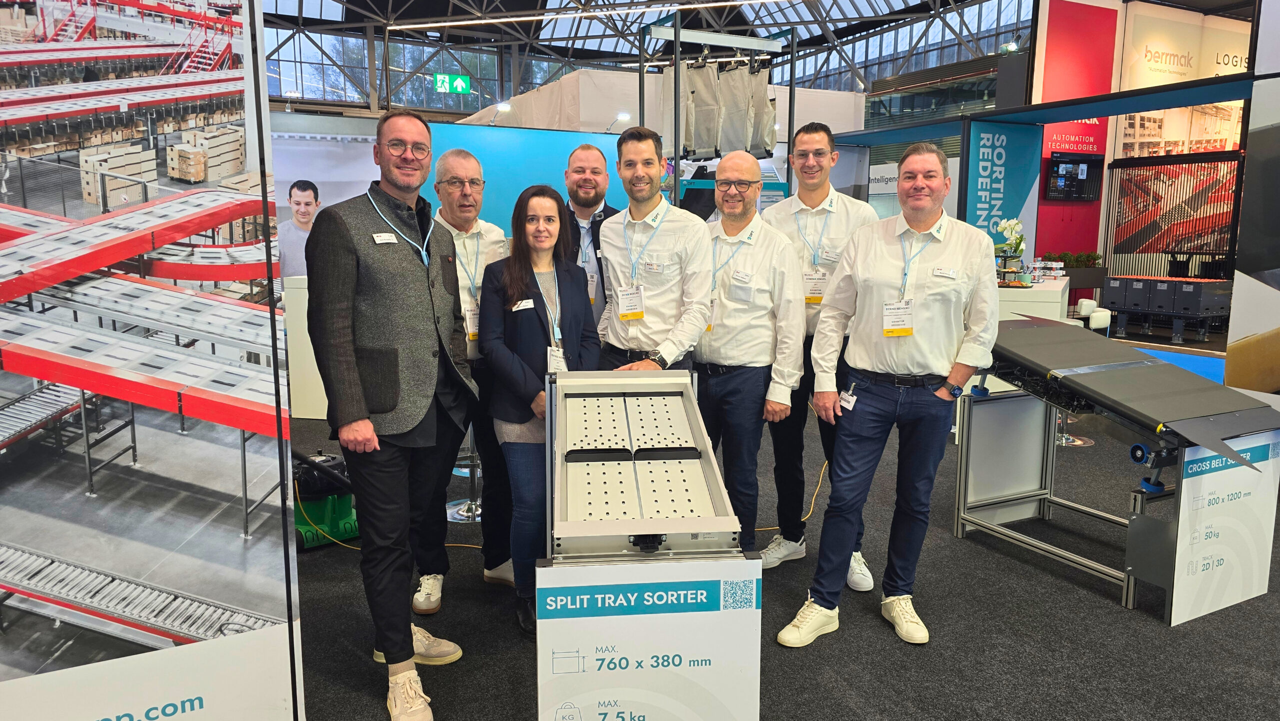 Eight people stand together smiling at an exhibition booth with display panels featuring conveyor systems, split tray sorters, and sorting equipment. Some wear name badges and lanyards, with machinery and display tables visible in the background.