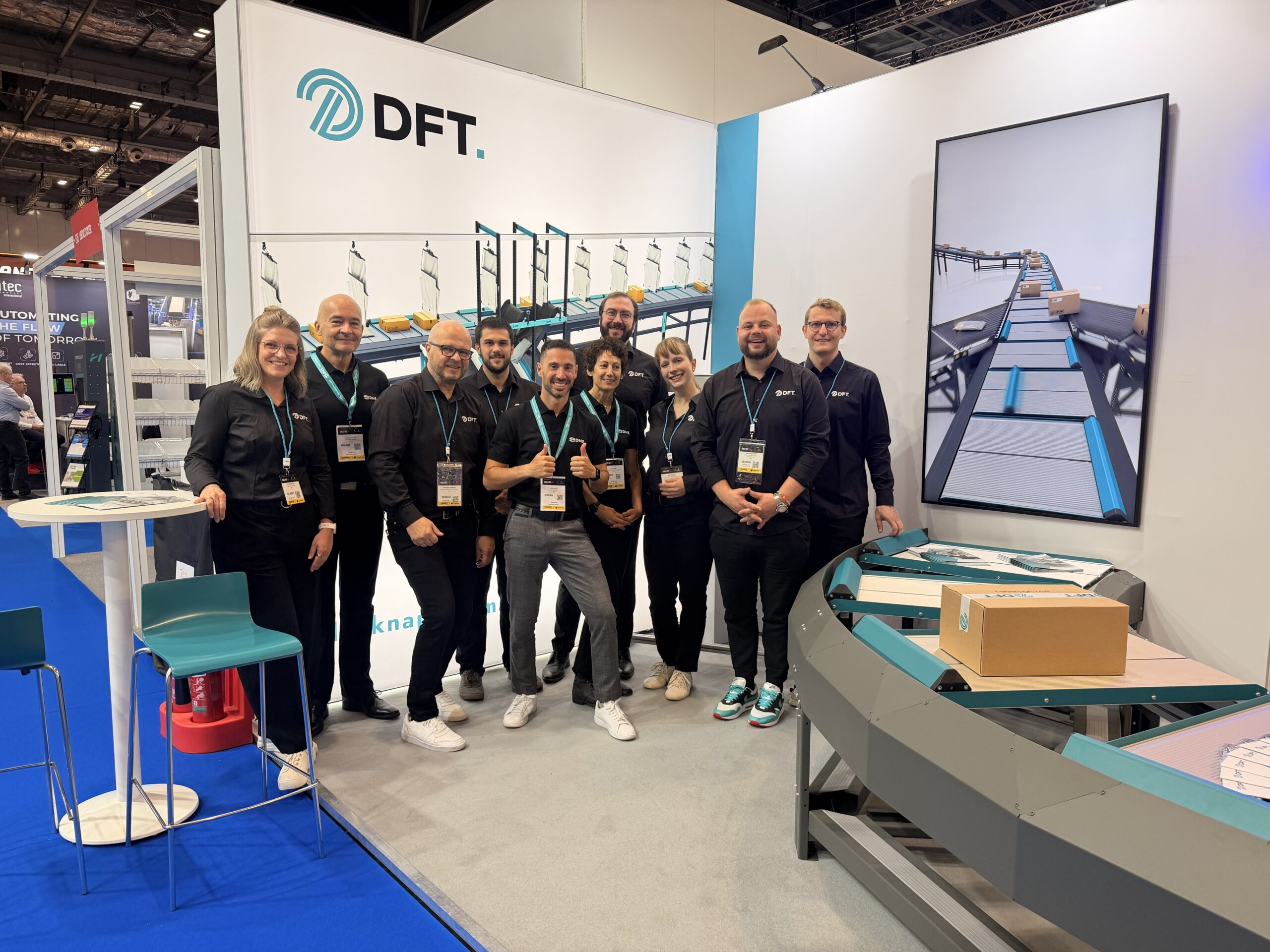Das Bild zeigt das DFT-Team, das an ihrem Messestand auf der Parcel + Post Expo 2026 posiert, mit einem Förderband und einer großen Visualisierung im Hintergrund.