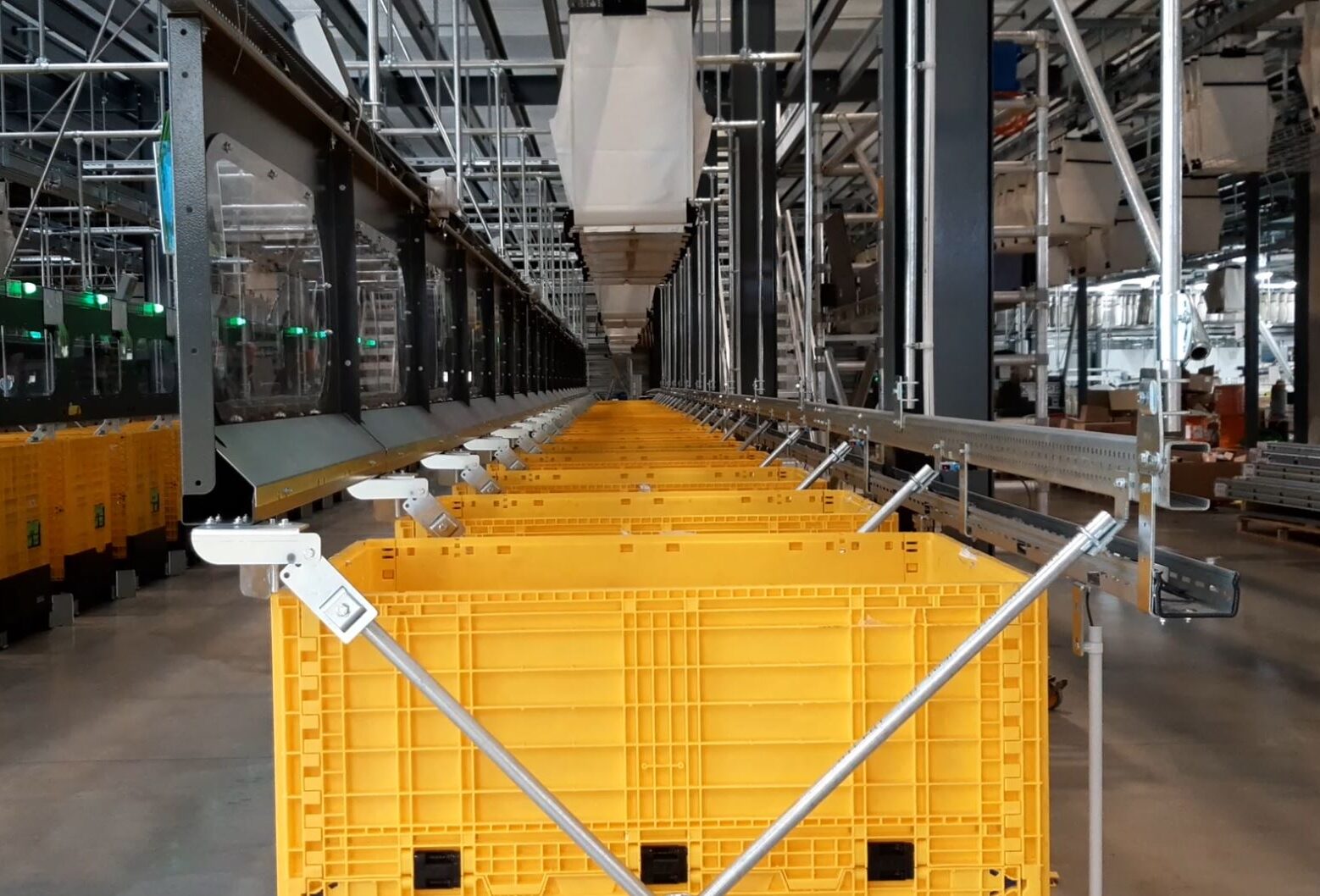 Blick auf ein automatisiertes Lagersystem mit einer Reihe gelber Behälter auf einem Förderband. Oben sind Metallträger und Maschinen zu sehen, die eine symmetrische industrielle Umgebung schaffen. Der Raum wirkt organisiert und effizient.
