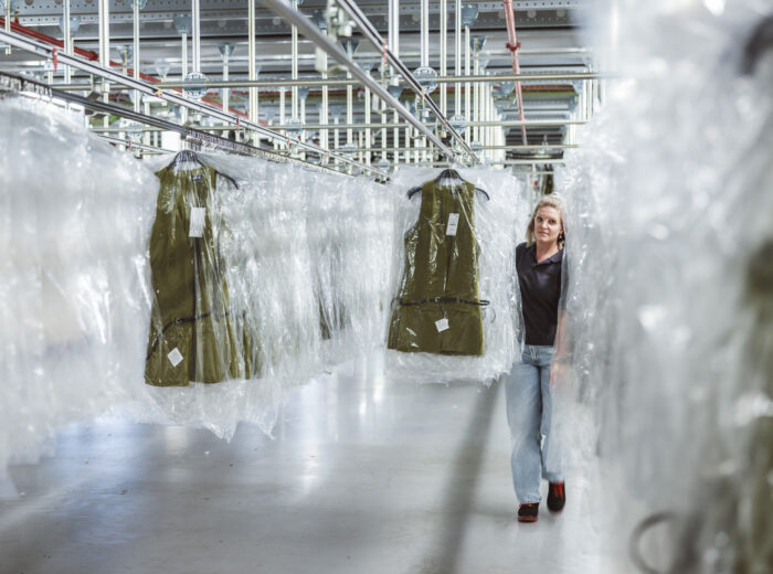 Eine Person geht durch ein großes Lagerhaus, das mit grünen Kleidungsstücken auf Bügeln gefüllt ist, die jeweils mit durchsichtigem Plastik abgedeckt sind. Der gut beleuchtete Raum ist mit Metallregalen ausgestattet, bereit, die Bestellungen des Onlinehandels effizient auszuführen.