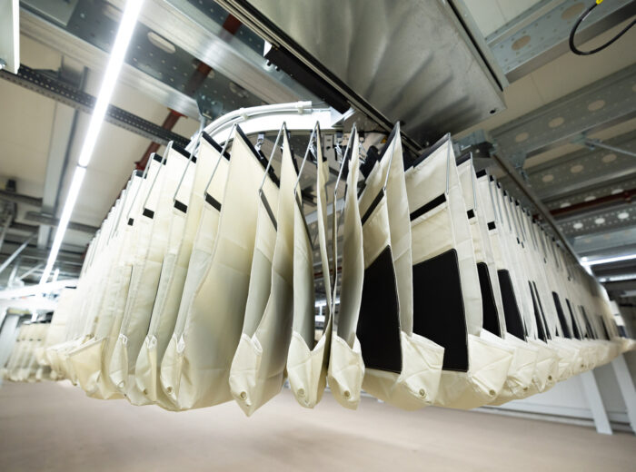 Reihen von beigen EcoPocket-Stoffbeuteln, die an einem automatischen Schienensystem in einer sauberen, industriellen Innenumgebung mit Metallträgern und heller Beleuchtung hängen.