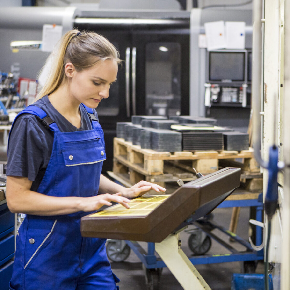Eine Frau in einem blauen Overall bedient ein Bedienfeld in einer Industriehalle, umgeben von Maschinen und Werkzeugen, die für die Ersatzteillogistik und den Maschinenbau unerlässlich sind, wobei im Hintergrund gestapelte Paletten zu sehen sind.