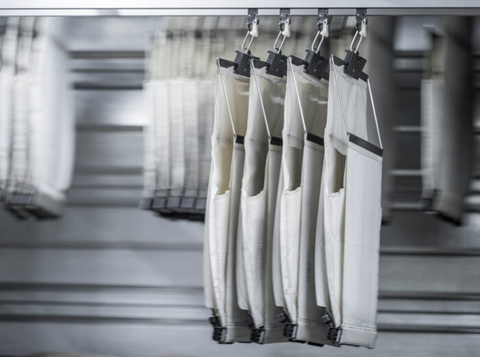 Vier weiße Industriefiltersäcke, die in einer Reihe an Klammern hängen, mit einem Metallhintergrund und weiteren unscharfen Filtersäcken im Hintergrund.