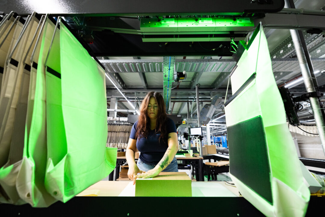 A woman with long hair works in a factory, assembling or inspecting a cardboard box under green-tinted industrial lights, surrounded by hanging fabric panels and machinery—an example of efficient Multi-Client Fulfillment in action.