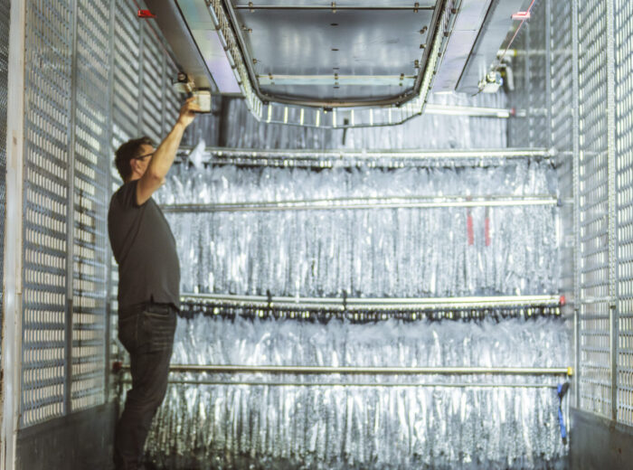 A person stands inside a large industrial space lined with silver metallic panels, inspecting a hanging conveyor overhead with one arm raised. The area appears to be a high-tech or specialized facility.