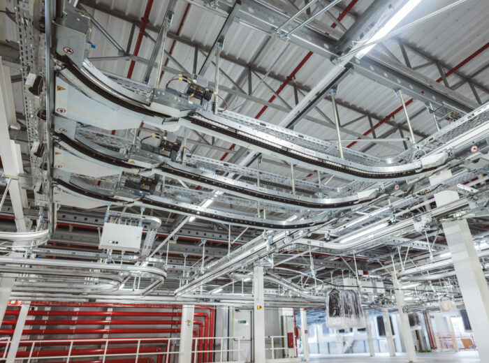 Ceiling view of an industrial facility showing a complex network of metal pipes, cable trays, and a hanging conveyor system, with bright lights illuminating the spacious, modern interior.