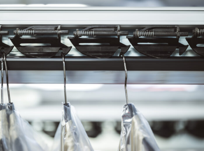 Close-up of a hanging conveyor system with clothing hangers holding plastic-covered clothes, likely in a dry cleaning or laundry facility. The focus is on the metal track and hooks.
