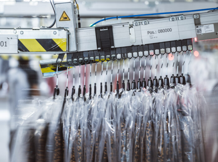 Clothes in plastic garment bags hang on a hanging conveyor system inside a modern industrial facility, possibly a dry cleaner or garment factory, with blurred background and machinery visible.