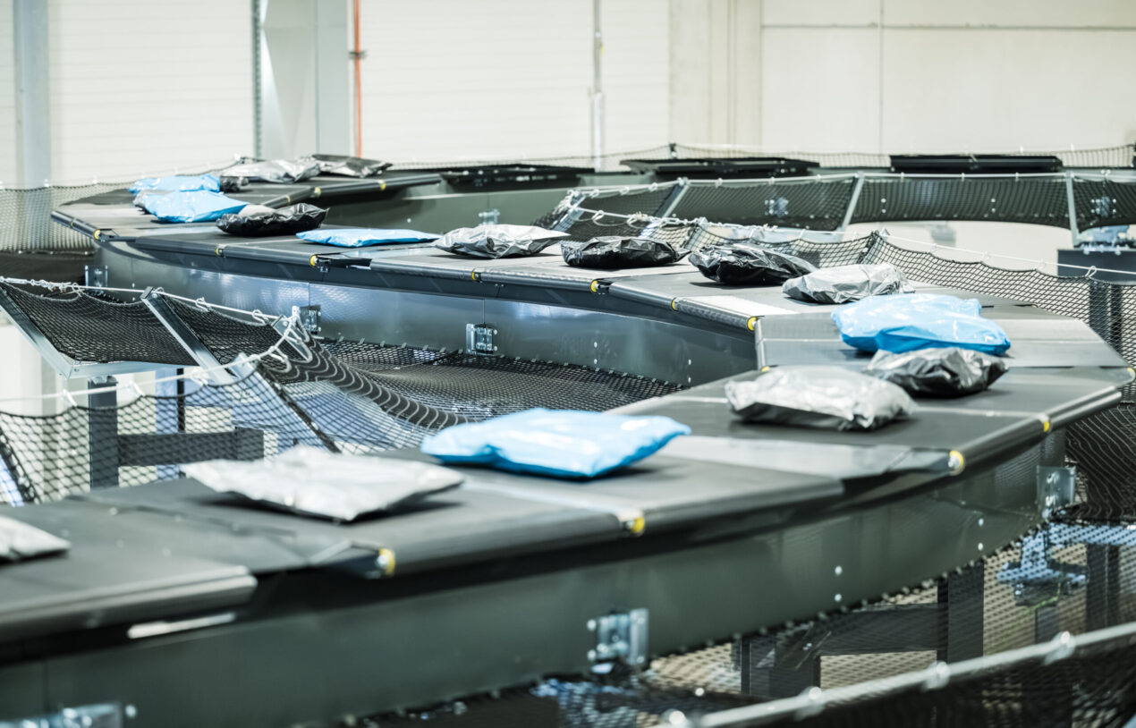 Ein Förderbandsystem in einem Lager, das von den Lösungen der LogiMAT inspiriert ist, transportiert Pakete, die in schwarze, blaue und graue Plastikfolie eingewickelt sind. Der Bereich wirkt organisiert und industriell und ist auf effiziente Sortierung und Auslieferung ausgelegt.