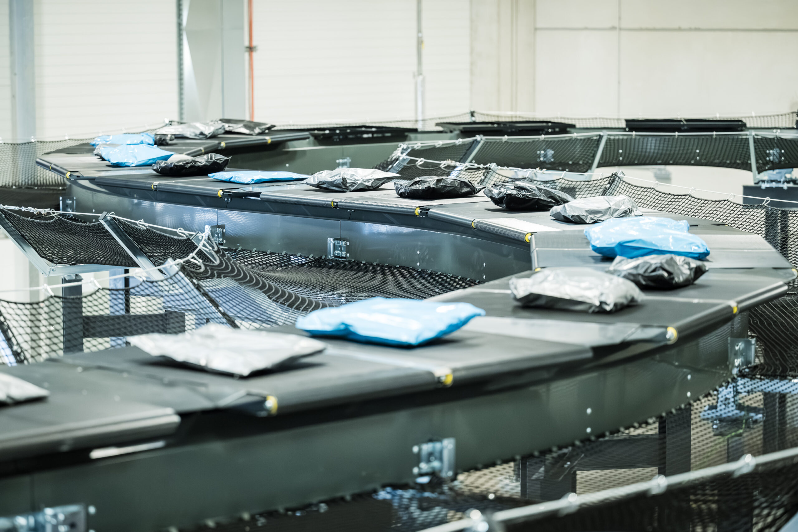 Ein Förderbandsystem in einem Lager, das von den Lösungen der LogiMAT inspiriert ist, transportiert Pakete, die in schwarze, blaue und graue Plastikfolie eingewickelt sind. Der Bereich wirkt organisiert und industriell und ist auf effiziente Sortierung und Auslieferung ausgelegt.