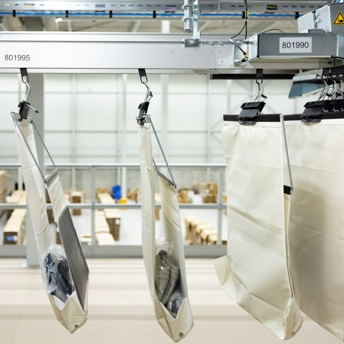 Ein Fördersystem mit hängenden Säcken, die verpackte Artikel enthalten, bewegt sich durch ein Industrielager, in dem Smart Sorting Solutions im Hintergrund mit Lagerbereichen und Regalen voller Kartons zu sehen ist.