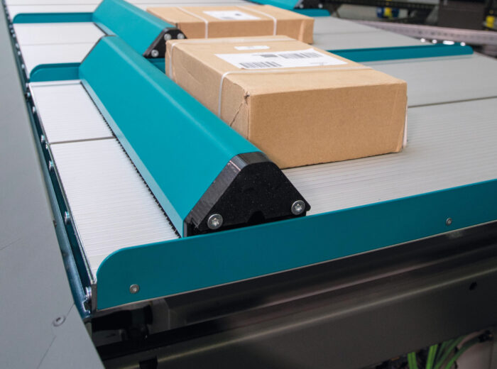 A brown cardboard box with a barcode label moves along a blue and white conveyor belt, directed by a Push Tray Sorter inside a modern industrial sorting facility.