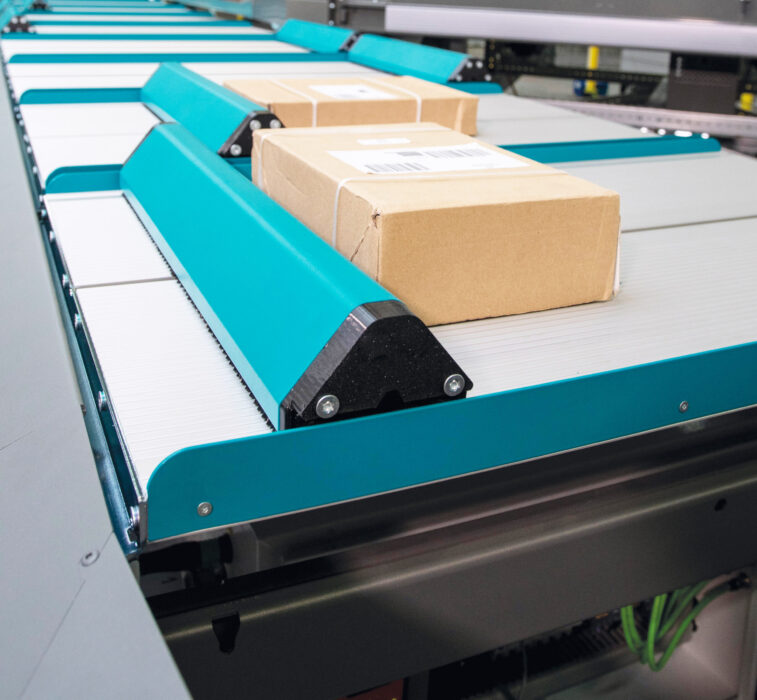 Ein braunes Kartonpaket mit einem Barcode-Etikett bewegt sich über ein modernes Push Tray Sorter-Förderbandsystem mit tealfarbenen und silbernen Komponenten in einem Lager oder Distributionszentrum.