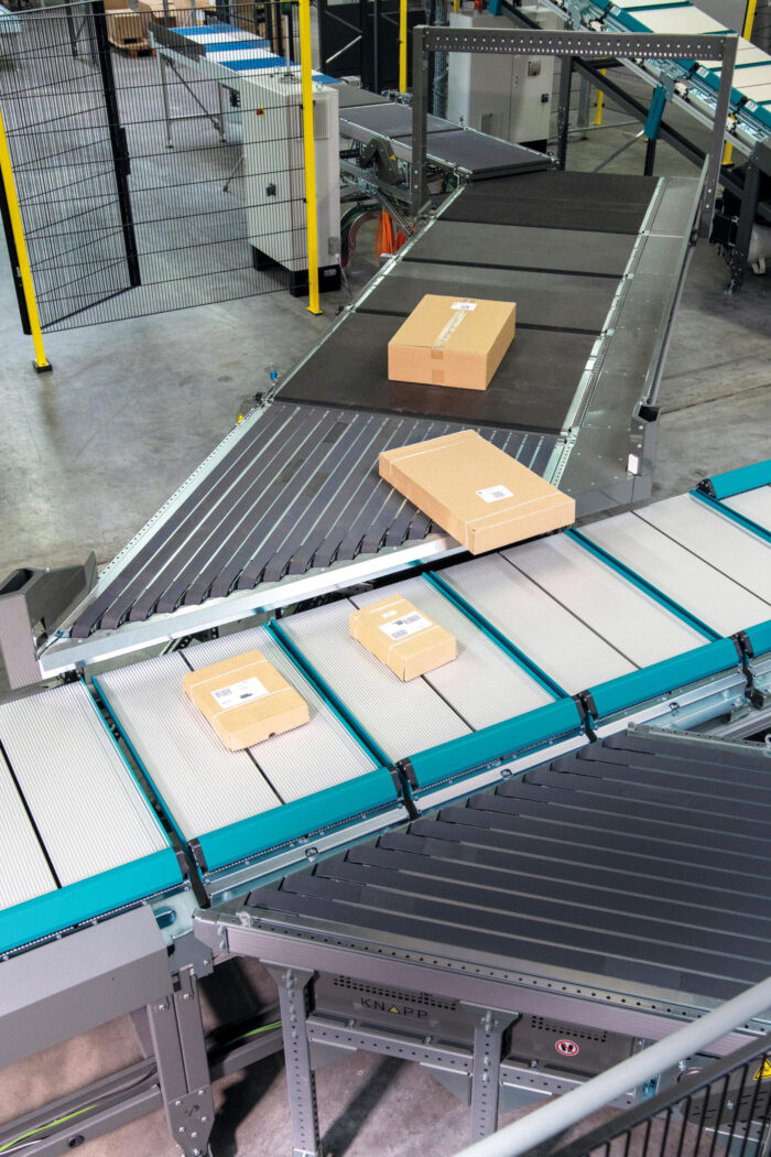Kartonverpackungen bewegen sich über ein Förderbandsystem in einem Lagerhaus. Im Hintergrund sind Industrieanlagen und Zäune zu sehen, während ein Push Tray Sorter die Sendungen effizient organisiert.