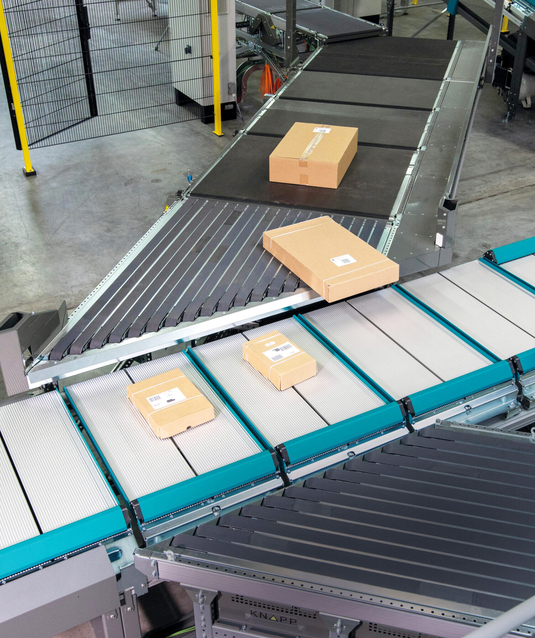 Several cardboard packages move along conveyor belts inside a warehouse, guided by a Push Tray Sorter for efficient sorting and shipment. The industrial surroundings feature metal fencing and advanced machinery.