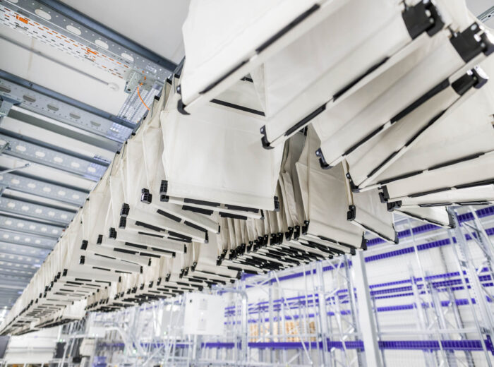 Rows of white fabric conveyor bags, part of a pocket sorter system, hang from metal rails in an automated warehouse or sorting facility, with shelves and storage racks visible in the background.