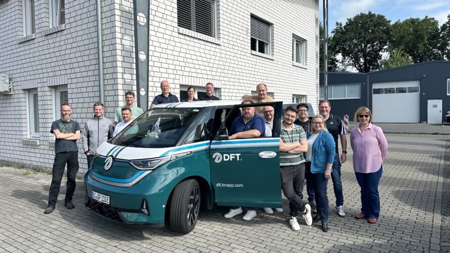 Eine Gruppe von Menschen steht an einem sonnigen Tag lächelnd neben einem blaugrünen und weißen Lieferwagen der DFT, der vor einem weißen Backsteingebäude geparkt ist. Die Tür des Lieferwagens ist offen, und einige Leute stehen im Inneren, während andere um ihn herum posieren.
