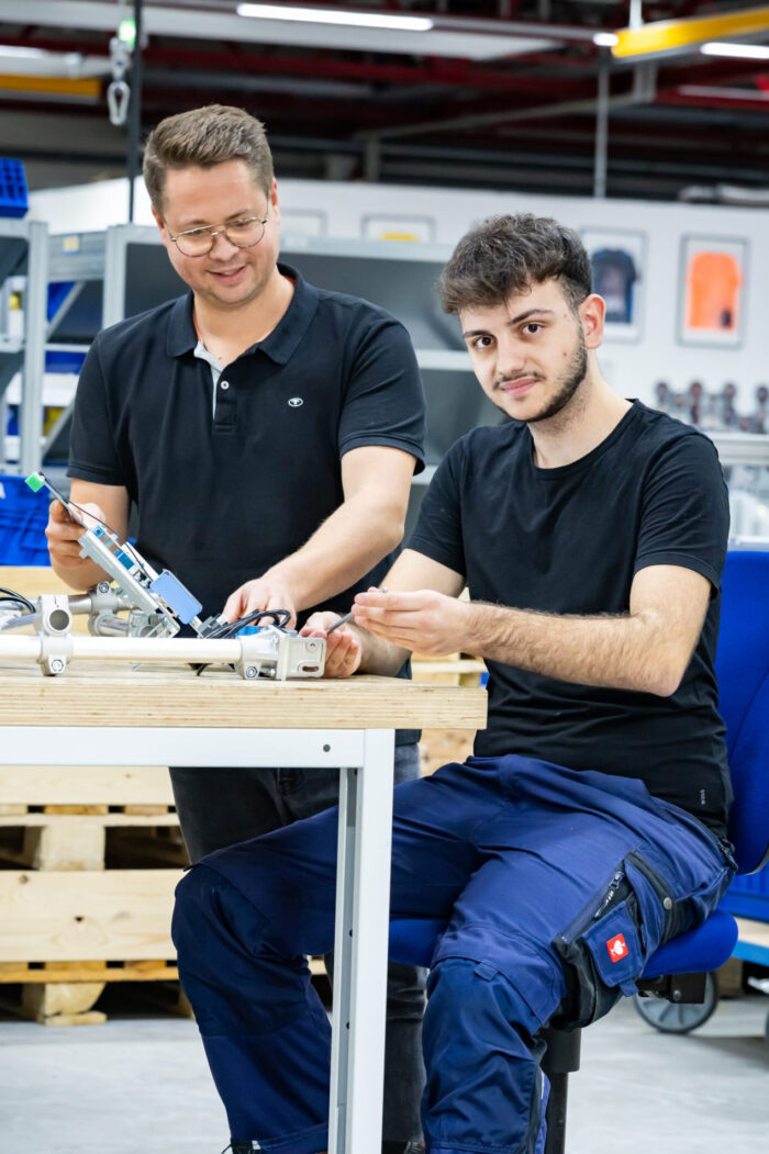 Zwei Männer arbeiten gemeinsam an einem Tisch mit elektronischen Bauteilen in einer Werkstatt; der eine sitzt und deutet auf das Gerät, der andere steht, lächelt und hält ein Werkzeug in der Hand – das Bild fängt eine praxisorientierte Ausbildungsumgebung mit Regalen und Geräten im Hintergrund ein.