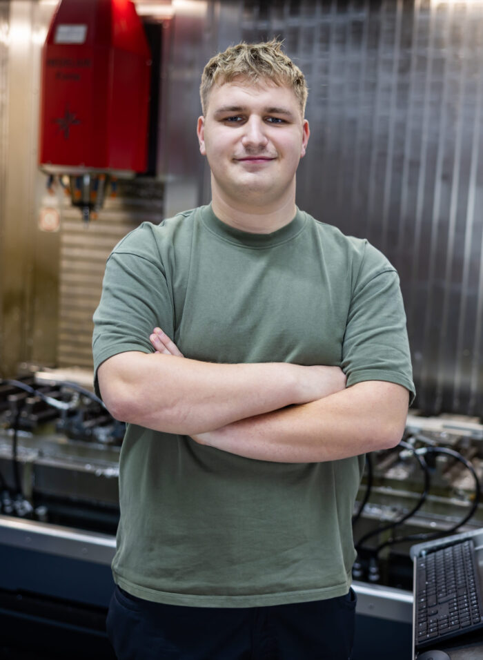 Ein junger Mann mit blonden Haaren, der ein grünes T-Shirt trägt, steht mit verschränkten Armen vor einer Industriemaschine in einer Werkstatt und ist bereit, seine Ausbildung zu beginnen.