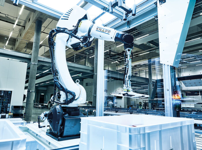 Ein Roboterarm in einer modernen Fabrik nimmt einen Gegenstand aus einem weißen Behälter. Die Umgebung ist mit Hightech-Maschinen und Industrieanlagen ausgestattet, was einen sauberen und organisierten Arbeitsplatz unterstreicht.