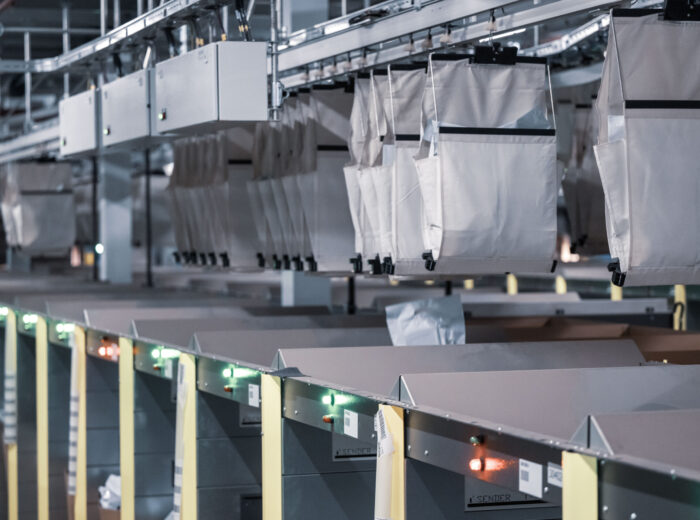 Fördersystem mit hängenden Stoffbeuteln in einer Lagerhalle. Die Beutel hängen an Schienen und bewegen sich in einer Reihe. Darunter befinden sich Regale mit Lichtern, die Sortiervorgänge anzeigen. Der Bereich ist schwach beleuchtet und wirkt industriell.