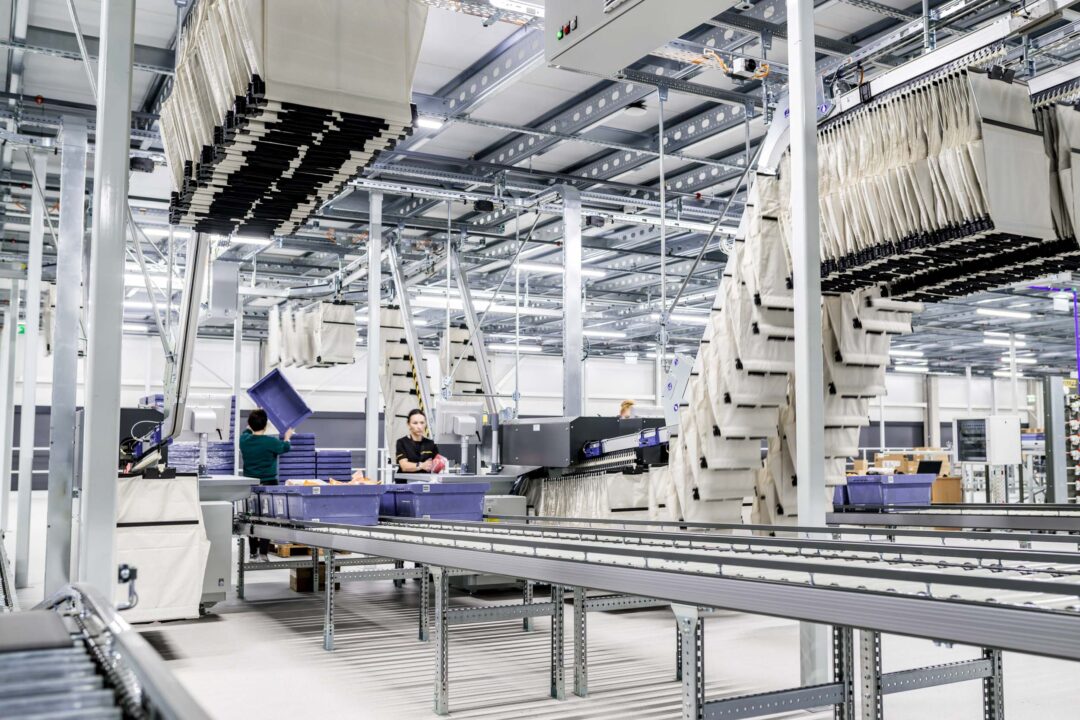 Das Innere eines modernen Lagers mit Förderbändern und automatisierten Systemen veranschaulicht die Effizienz der Pharmalogistik. Arbeiter handhaben blaue Behälter, während die hohe Decke mit sichtbaren Metallträgern für Geräumigkeit sorgt. Stapel von Artikeln stehen in einer hellen, gut beleuchteten Umgebung.
