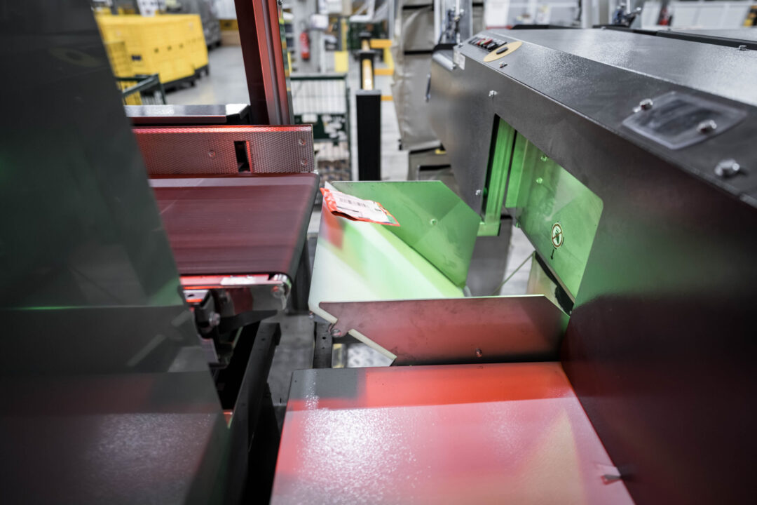 Sorting machine processing a small package on a conveyor belt inside an industrial facility. The package is moving under a scanner with green and red lights. Yellow storage bins are visible in the background.