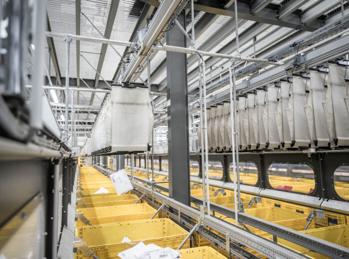 A warehouse interior with a system of conveyor belts and yellow bins arranged below. Fabric chutes hang from above, guiding parcels into the bins. The scene is industrial and organized, suggesting an automated packaging or sorting process.