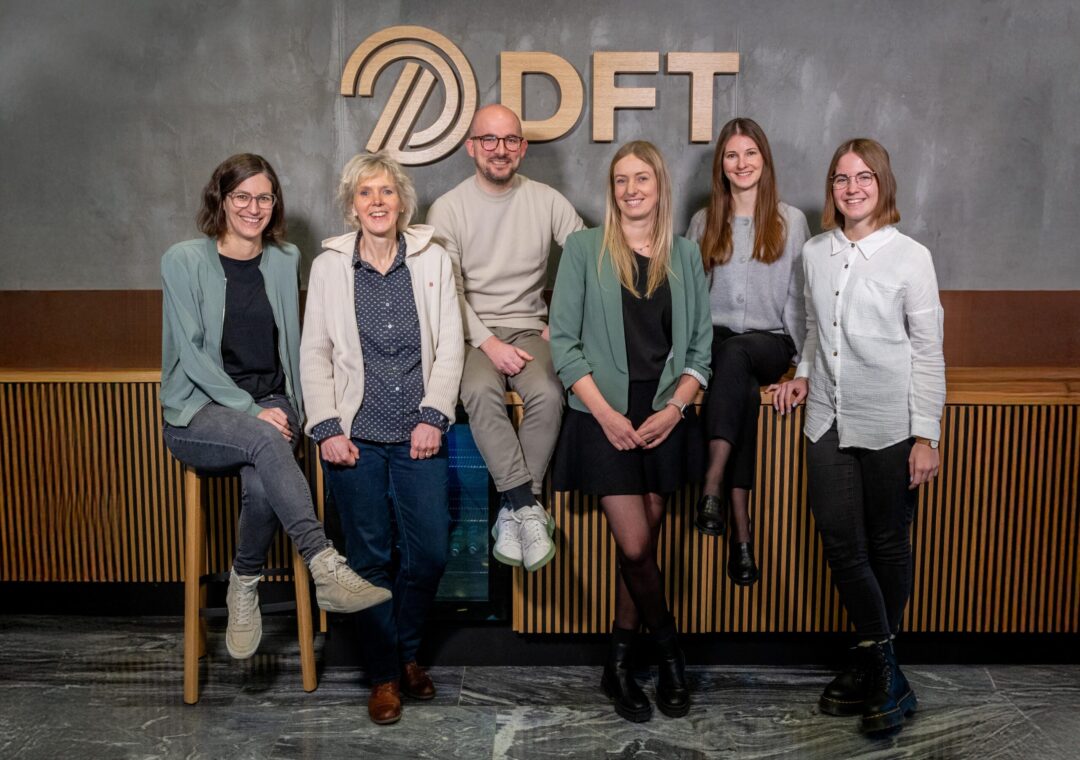 A group of six people, three men and three women, are smiling and posing in front of a wall with the letters DFT. They are casually dressed, and some are sitting or leaning against a wooden bench.