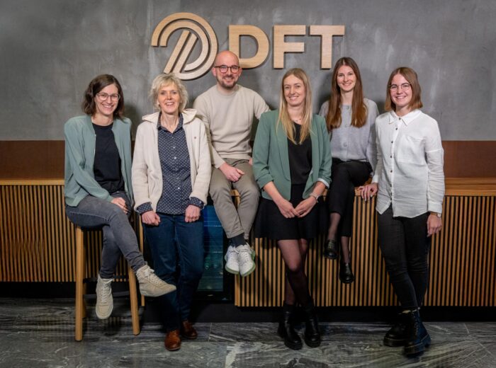 A group of six people, three men and three women, are smiling and posing in front of a wall with the letters DFT. They are casually dressed, and some are sitting or leaning against a wooden bench.